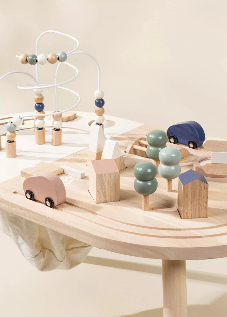 A variety of wooden toys on a Wooden Activity Table, including toy cars, houses, and a bead maze, all in pastel colors—blue, pink, and cream—set against a neutral background.