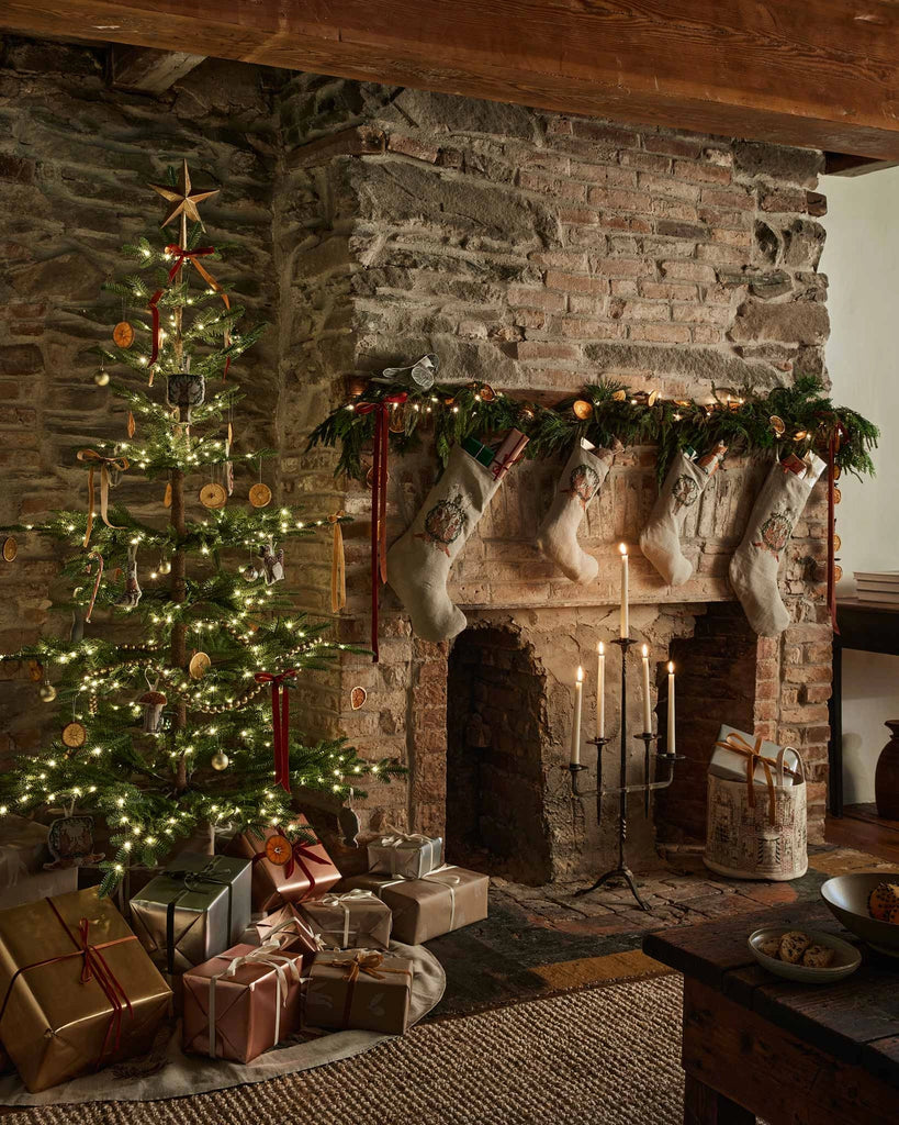 A cozy stone fireplace with stockings and garland sits beside a lit Christmas tree decorated with ornaments, including the Coral & Tusk Togetherness Deer Ornament. Wrapped gifts beneath the tree complete this festive holiday scene.