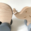 Two wooden toy elephants are linked together by their trunks, creating an adorable Wooden Mom and Baby Elephant Push Toy Set. Each has gray wheels and is crafted from light-colored wood with simple facial features. Perfect for developing fine motor skills, the toys are free of harmful chemicals. The background is plain white.