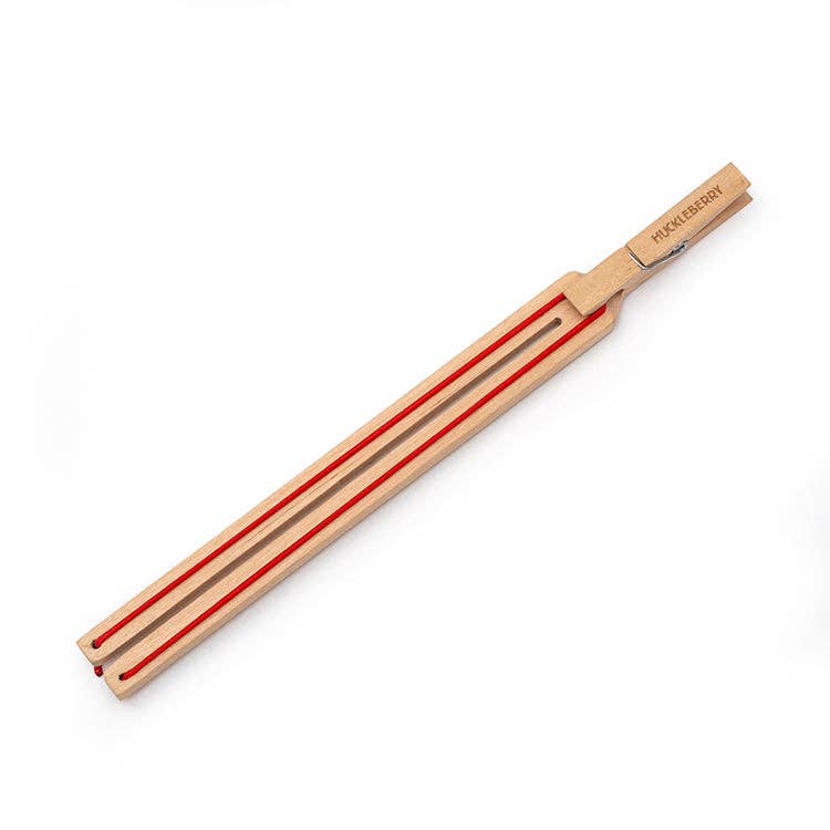 The Huckleberry Paper Plane Launcher is a birch wood tool with two prongs and a flat handle, accented by red grooves along the prongs, designed to resemble a paper airplane in flight.