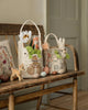 The Coral & Tusk Easter Parade Basket, decorated with animal and character designs, sits on a wooden bench filled with plush toys, flowers, and greenery, surrounded by colorful eggs and a small toy deer.