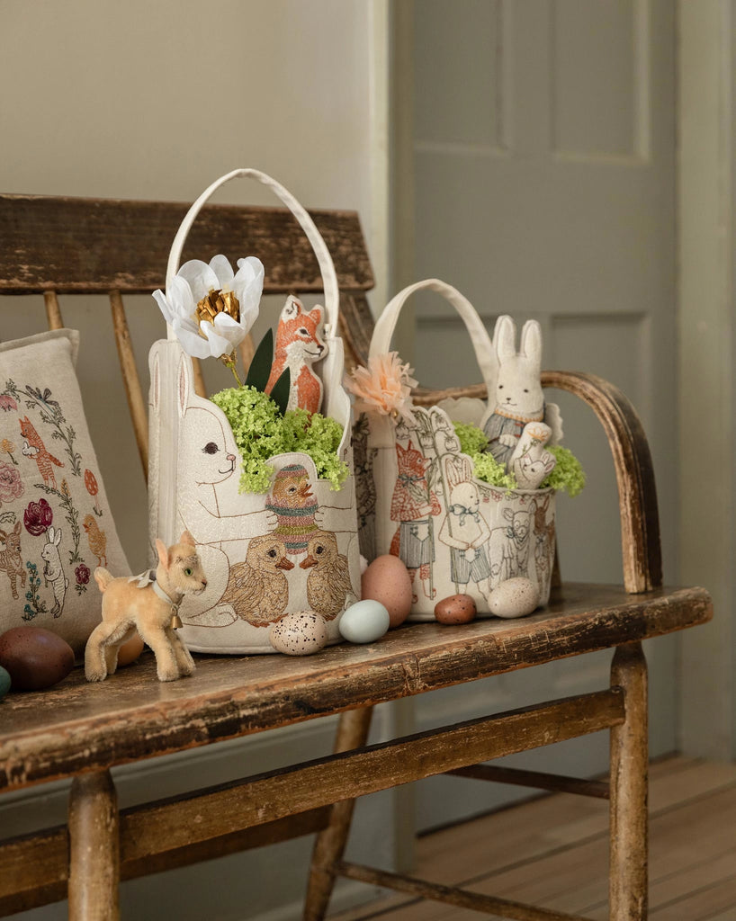 The Coral & Tusk Easter Parade Basket, decorated with animal and character designs, sits on a wooden bench filled with plush toys, flowers, and greenery, surrounded by colorful eggs and a small toy deer.