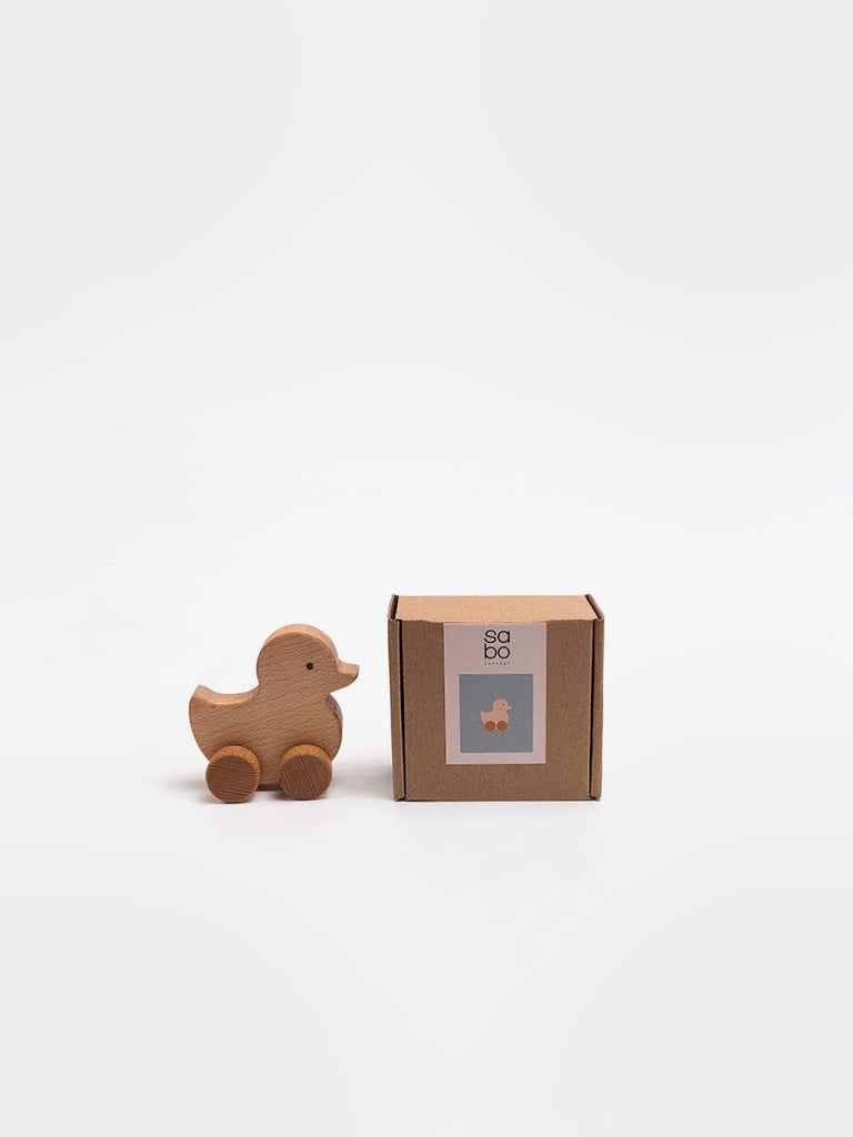 The Sabo Concept Rolling Duck - Natural, a wooden duck toy with wheels crafted from natural materials in the SABO concept style, is positioned beside a brown cardboard box that displays a small image of the same toy on its label. Both items are set against a plain white background.