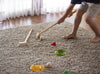A child plays indoor mini golf on a beige carpet, using the Mini Golf Set's clubs to putt balls toward target holes marked with colorful flags, while sunlight filters through sheer curtains in the background.
