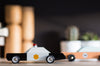 The Candylab Police Cruiser, crafted from solid beech wood, showcases a black and white body adorned with a yellow star emblem on the side. It features rubber tires and a red light on top. Safety tested for durability, the wooden toy police car rests on a table amidst additional toys and slightly blurred household items in the background.