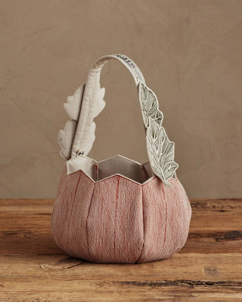 The Coral & Tusk Jack-o'-Lantern Basket features a soft, pleated fabric body in muted pink tones with a handle detailed in white leaves, blending style and Halloween charm. Ships in one week. Displayed on wood against a beige background.