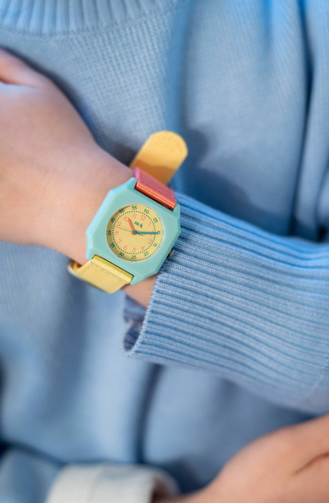 A child wears a pastel blue sweater and the Mini Kyomo Watch - Bubble Gum, featuring a blue face, pink and yellow straps, and a cream numbered dial—an ideal playful kids watch.