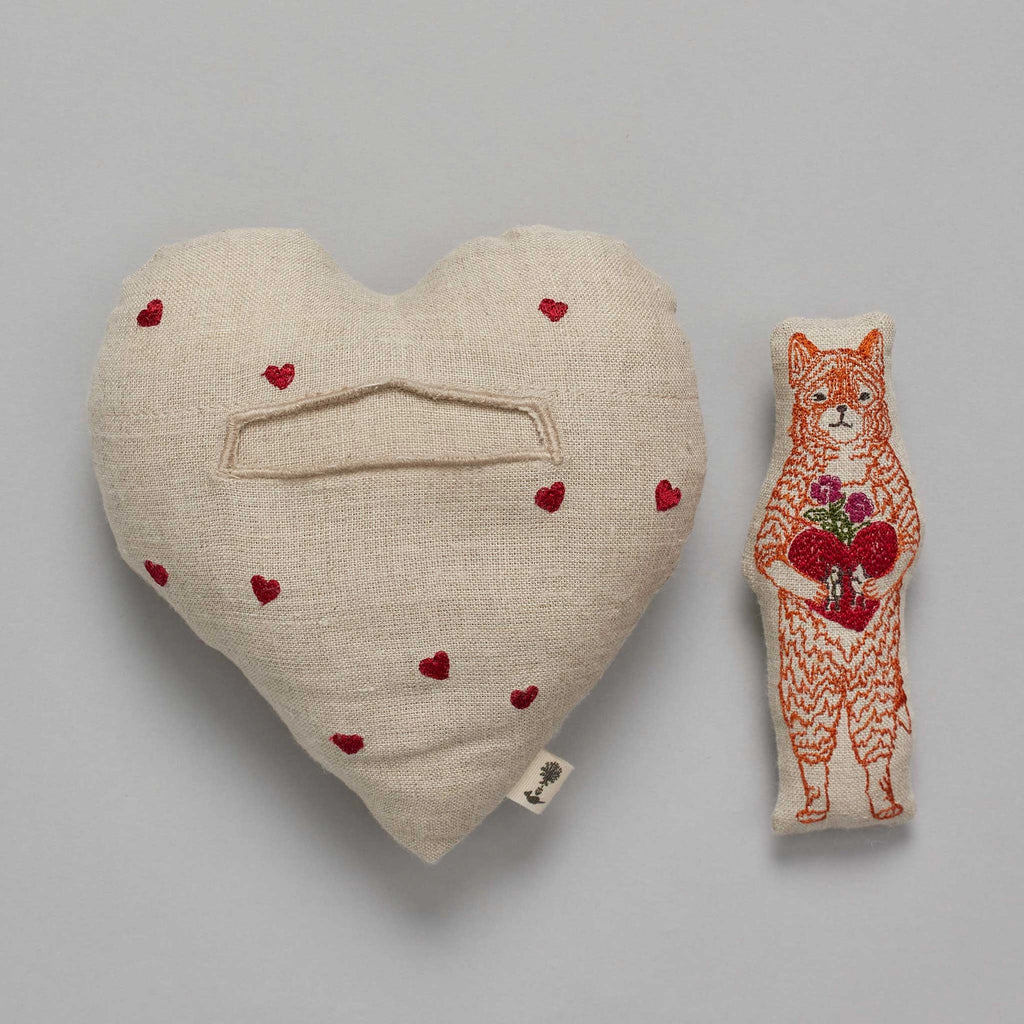 A Coral & Tusk Fox Heart Pocket Valentine, adorned with small red hearts and featuring a fabric bookmark shaped like a fox holding an embroidered heart, all on a bed.