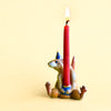 A Mouse Cake Topper sitting upright, painted in light brown with blue accents, holds a lit red candle on its back against a plain yellow background.