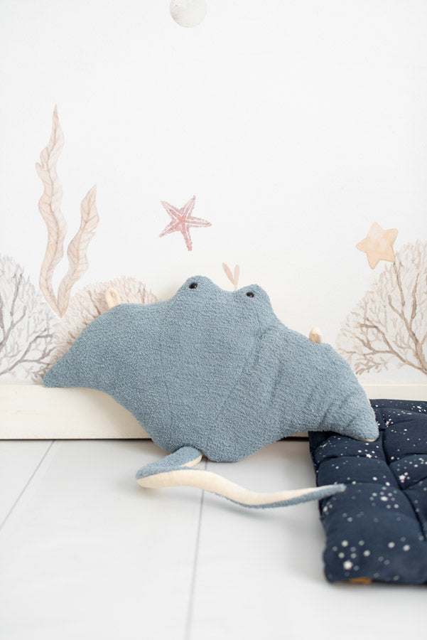 A plush Light Blue Stingray toy with a light blue texture, prominent eyes, and a tail, set against a simple backdrop featuring drawn seaweed and starfish.