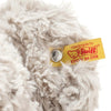 A close-up of the Steiff, Honey Teddy Bear showcases its fluffy, light-colored fur. Attached to the toy's ear is a yellow tag featuring a red teddy bear logo and the text "Handmade by Steiff," along with a signature metal button, indicating it is part of the Soft Cuddly Friends Collection.