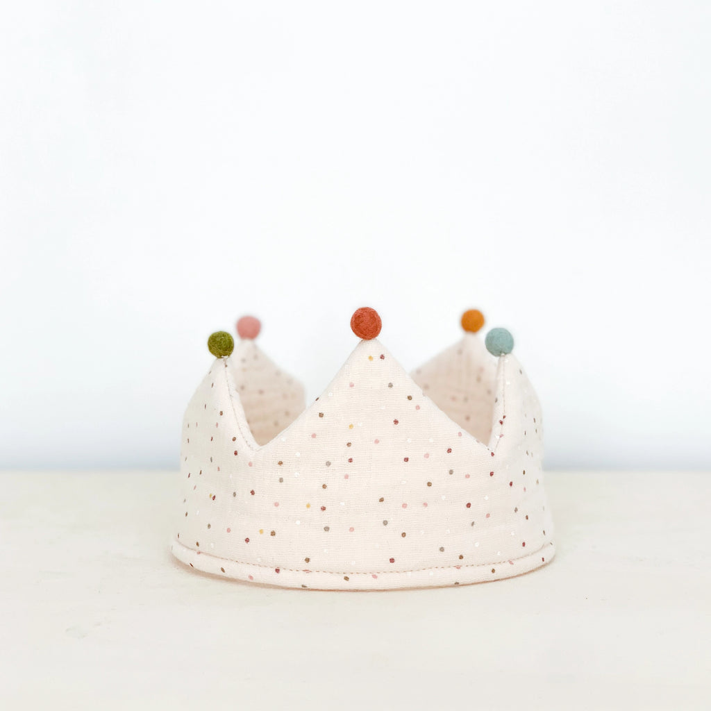 A soft, fabric Confetti Reversible Birthday Crown with a white base and small, multicolored dots. This handmade crown features five triangular points, each topped with a different colored button: green, orange, blue, and yellow. Made of organic cotton, it boasts a clean, light gray background.