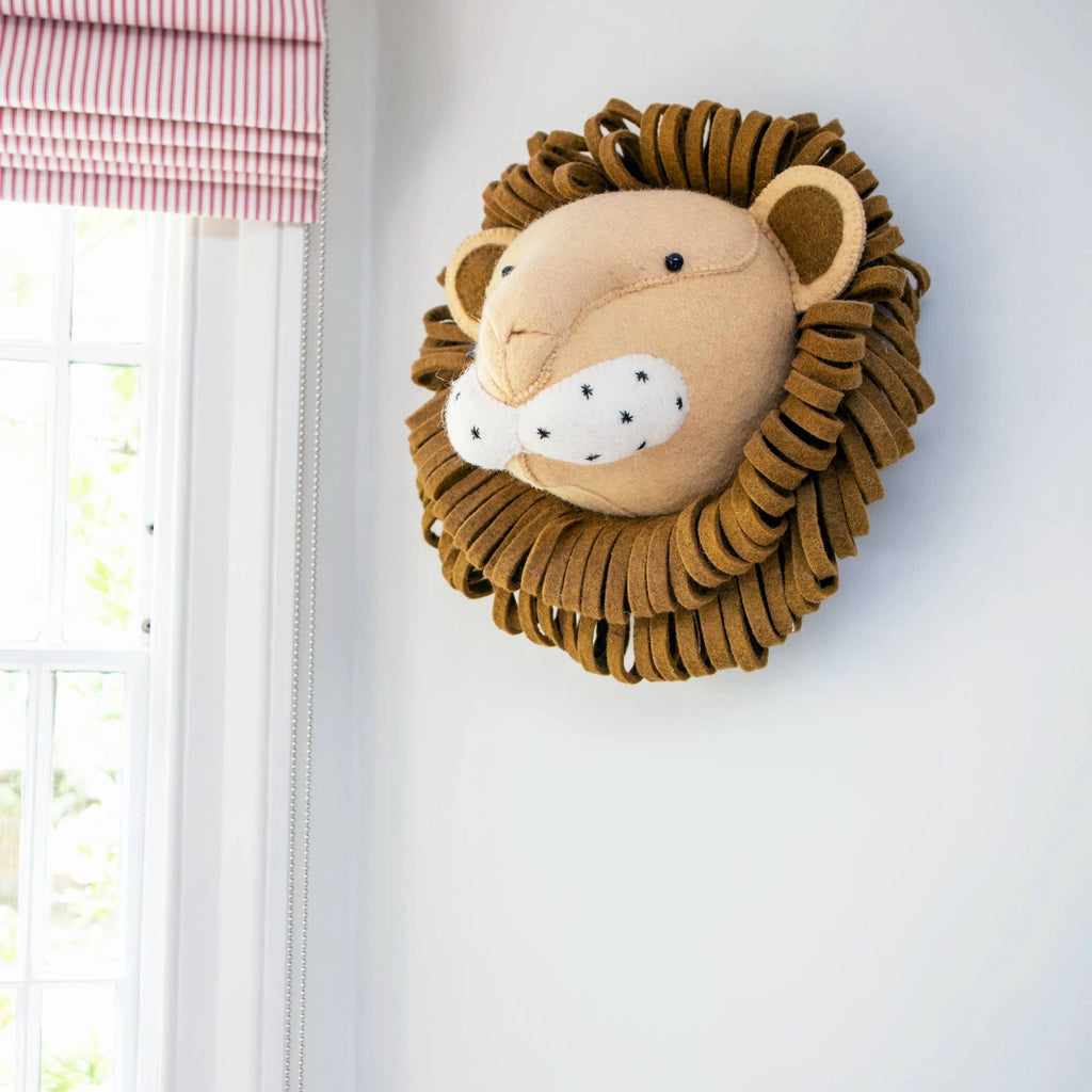 A handcrafted felt lion wall decor - large with a friendly face and layered fabric mane, mounted on a white wall beside a window with a red and white striped valance.