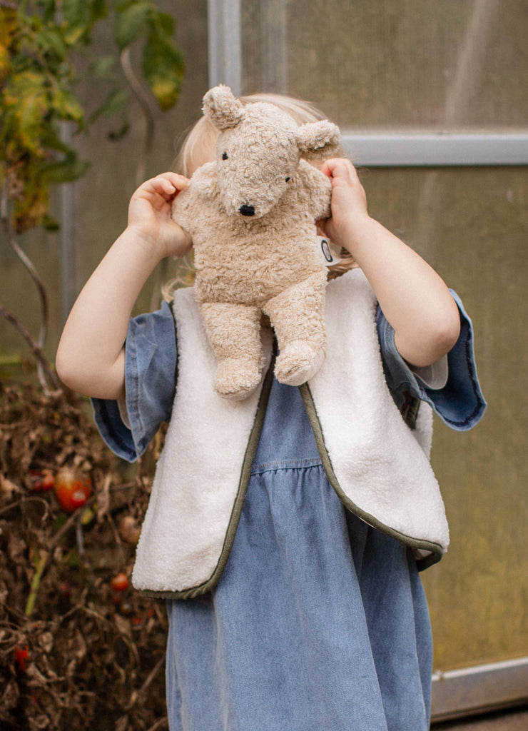 A young child in a blue dress holds a Senger Naturwelt Cuddly Animal - Linen Bear in front of their face, obscuring it, while standing outdoors near plants and a glass door.