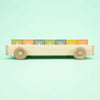 A handmade wooden toy car with four round wheels is positioned side-on against a light green background. Six colorful Uncle Goose Classic ABC Blocks with Wagon sit inside the basswood wagon, each block displaying different letters in various fonts and decorative patterns.