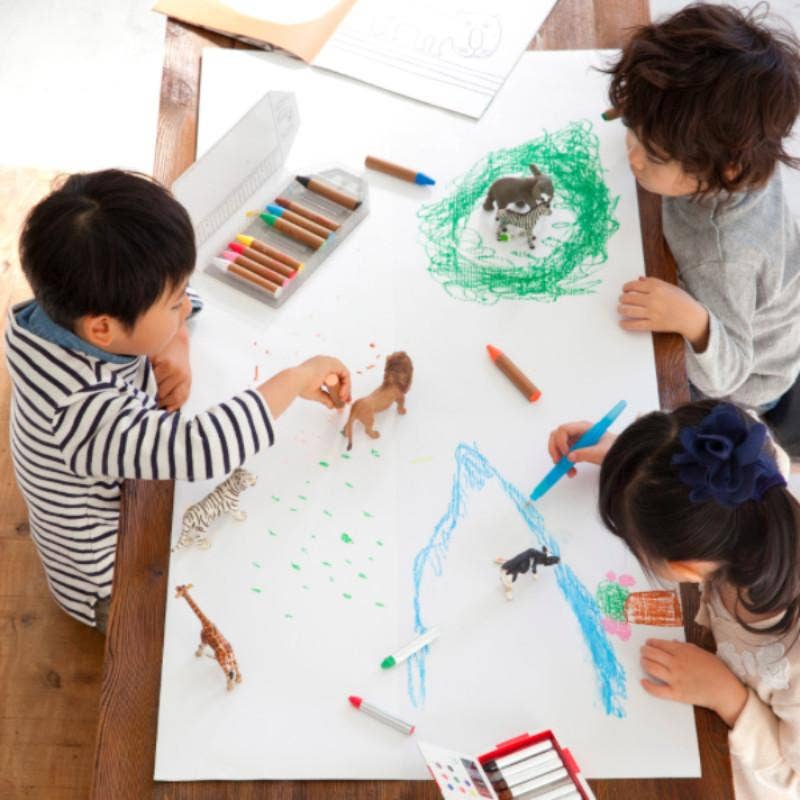 Three children are working on a large sheet of paper, using colorful markers to create their artwork. They are surrounded by toy animals such as a giraffe, lion, and zebra. Kitpas Crayons - 6 Colors contribute vibrant touches to their creations. Other non-toxic drawing tools are also available in this bright and imaginative setting.