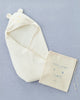 A cream-colored, organic cotton Baby Swaddle with a hood featuring bear ears and an embroidered bear face is spread on a light blue surface. Next to it, there is a drawstring pouch with the text "hooded swaddle" and a cute animal face illustration. Perfect for 0-3 months.