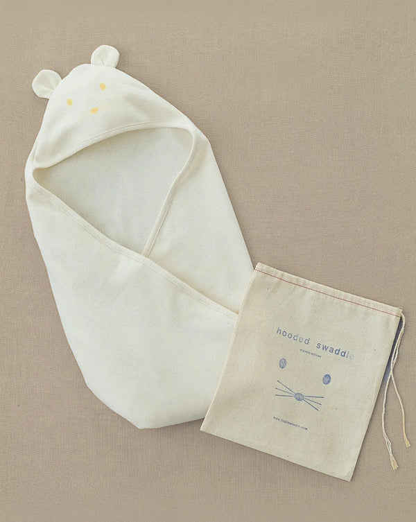 A cream-colored Baby Swaddle made from organic cotton, adorned with small ears, is displayed on a neutral backdrop. Accompanying it is a drawstring pouch labeled "hooded swaddle," showcasing a minimalist animal face design—ideal for newborns or as an ideal gift for baby showers.