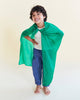 A young child smiles barefoot in a white shirt, blue pants, and an Emerald Sarah's Silks Solid Color Playsilk cape, striking a playful superhero pose perfect for open-ended play against a plain light background.