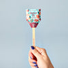 A hand with blue-painted nails holds a Pollinator Seed Pop (Single), decorated with colorful floral designs and "Plant Me!" text. Packed with wildflower seeds, it's perfect for starting your pollinator garden. The plain background is light blue.