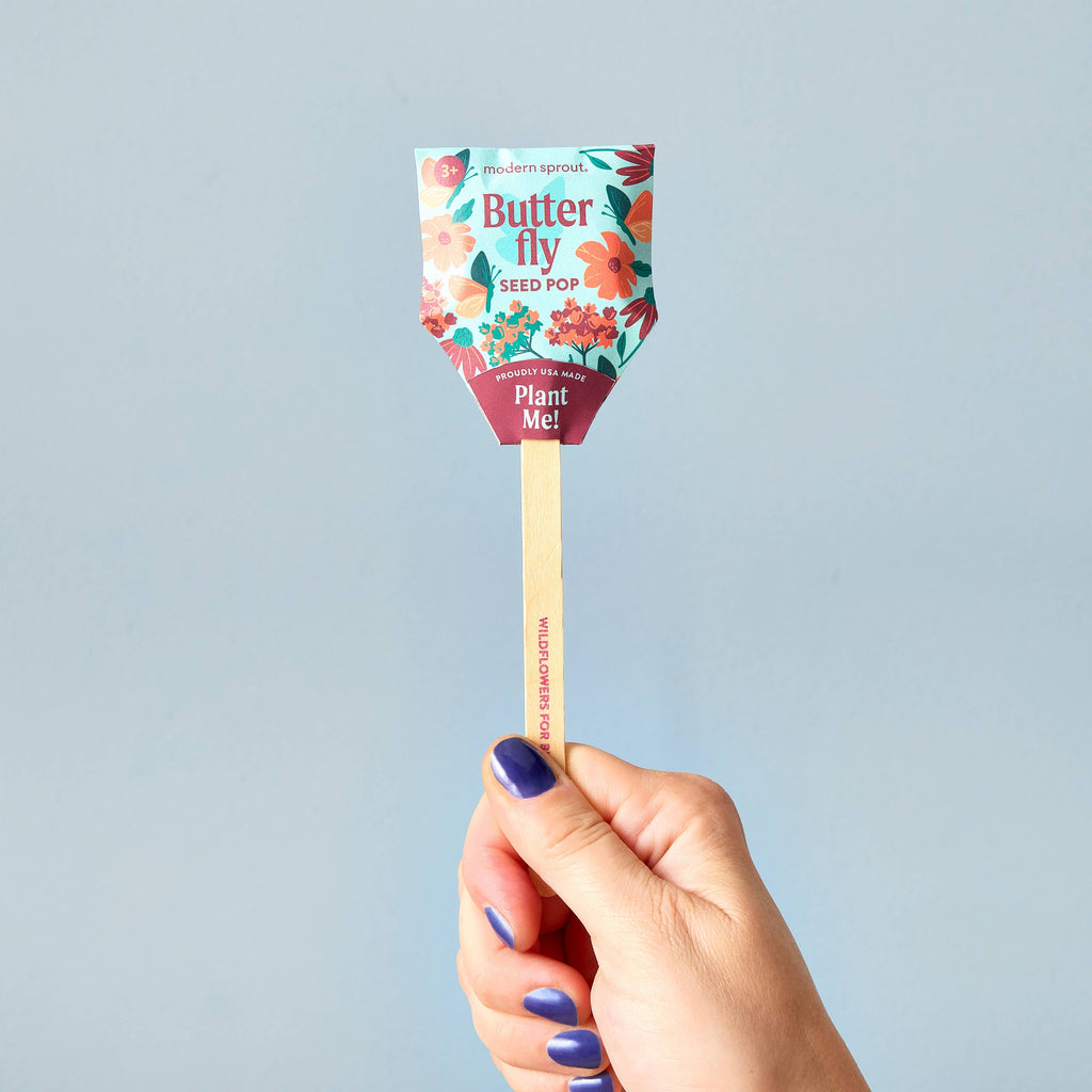 A hand with blue-painted nails holds a Pollinator Seed Pop (Single), decorated with colorful floral designs and "Plant Me!" text. Packed with wildflower seeds, it's perfect for starting your pollinator garden. The plain background is light blue.