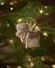 The Coral & Tusk Reindeer with Bells Ornament, featuring antlers, a red beaded necklace, and golden bells, hangs on a Christmas tree branch illuminated by warm white string lights.
