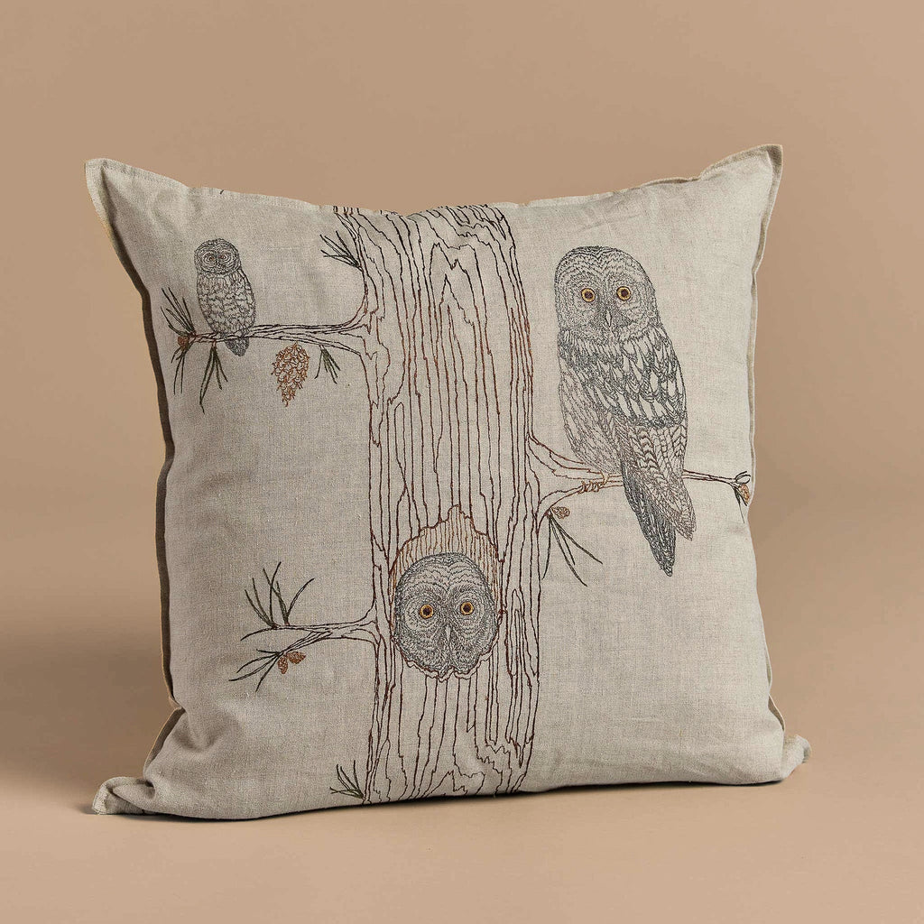 Discover the Coral & Tusk Owl Family Tree Pillow: a beige embroidered linen piece featuring a tree adorned with three owls. One perches among pine cones, another rests in a hollow, and one graces the trunk against a neutral brown backdrop.