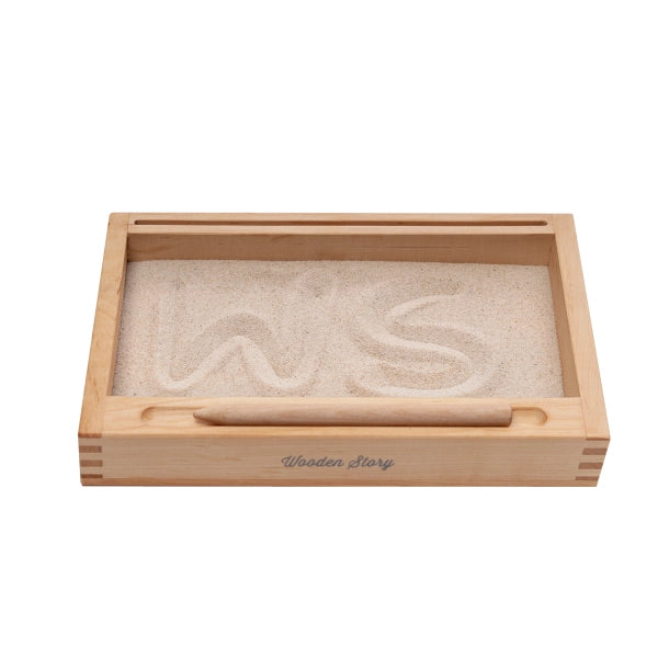 The Sand Tray with Flash-Card Holder is a rectangular wooden Montessori tray filled with fine sand, perfect for developing writing skills. The letters "WS" are drawn in the sand. A wooden stick rests on the front edge of the tray, which is labeled "Wooden Story." This educational toy combines learning and play beautifully.