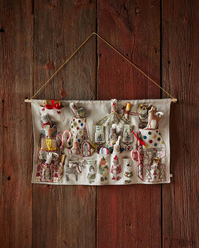 The Coral & Tusk North Pole Advent Calendar is a festive fabric calendar featuring stitched holiday scenes, small pockets for ornaments, candy canes, and mini characters, hanging from a wooden wall by a string.