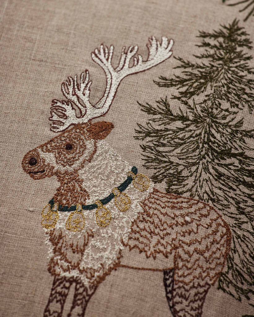 An embroidered reindeer wearing a bell collar stands next to an evergreen tree on this Coral & Tusk Christmas Cheer Lumbar Pillow.