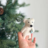 A hand holds a small, white Polar Bear Finger Puppet: Polar Bear Catching a Fish ornament in front of a green Christmas tree—a charming addition for imaginative play and Arctic storytelling this holiday season.
