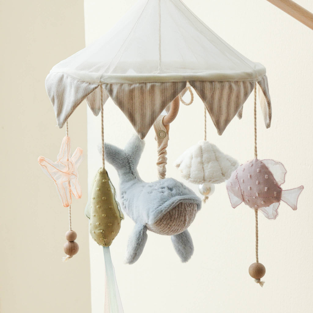 The Nautical Dreams Mobile hangs from the ceiling with plush sea shapes—whale, shell, fish, and star. Neutral tones with pastel accents and wood beads make it perfect nursery decor.