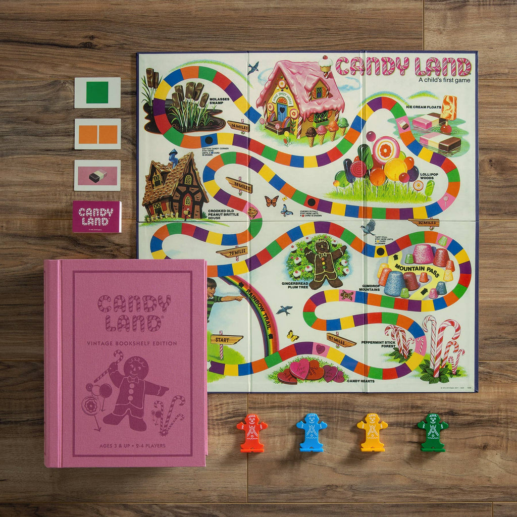 The Board Game: Candy Land Vintage Bookshelf Edition is displayed on a wooden surface, featuring its colorful board, four colored playing cards, a pink vintage bookshelf box, and gingerbread player pieces in green, yellow, blue, and orange.