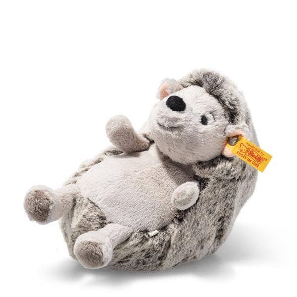 The Steiff Hedgy Hedgehog 6" is a plush toy with soft gray and white fur, a smiling face, small ears, a round nose, and a yellow ear tag. It lies on its back against a plain white background.