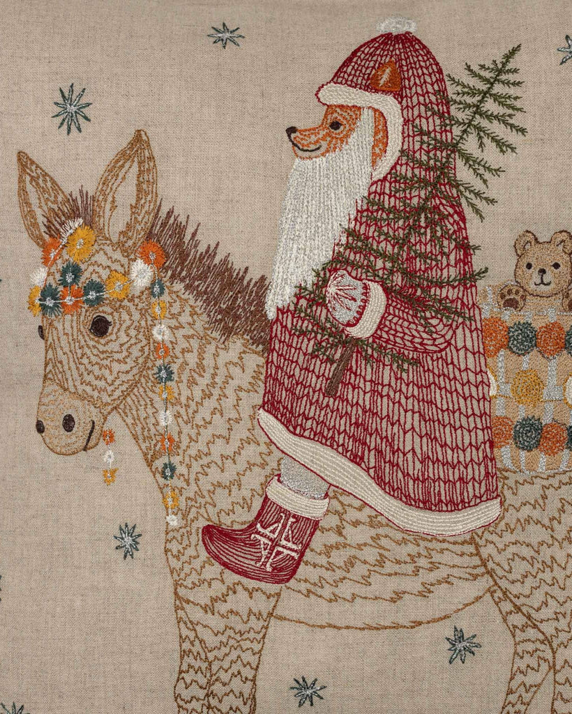 The Christmas Donkey Pillow features a festive donkey with a floral crown, carrying a fox dressed as Santa and a teddy bear. The fox holds a tree amid sparkling blue stars—perfect for adding holiday cheer to your décor.