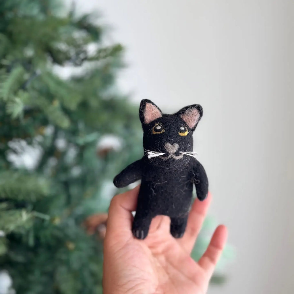 A hand holds a small Black Cat Finger Puppet, needle-felted with white whiskers and pink inner ears. Crafted from 100% wool, this adorable handmade plushie stands out against the backdrop of a green, blurry pine tree.