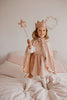 A young girl stands on a bed, smiling in a soft pink dress and white knee-high socks. She wears the Cape - Pink sequins with a sparkly crown and holds a star wand. The room has light bedding and decor.