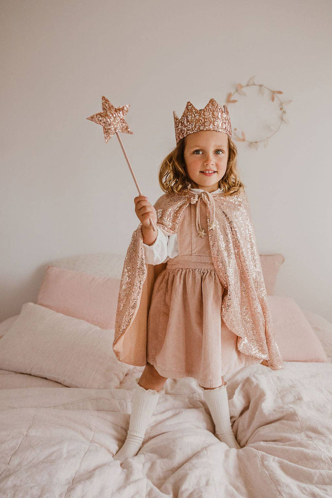 A young girl stands on a bed, smiling in a soft pink dress and white knee-high socks. She wears the Cape - Pink sequins with a sparkly crown and holds a star wand. The room has light bedding and decor.
