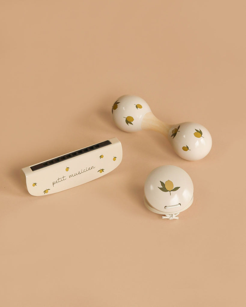 The Konges Sloejd Petit Music Set - Glossy Lemon, featuring three small cream-colored wooden instruments with yellow lemon patterns—a harmonica, kids’ maracas, and a jingle bell—rests on a light beige background.