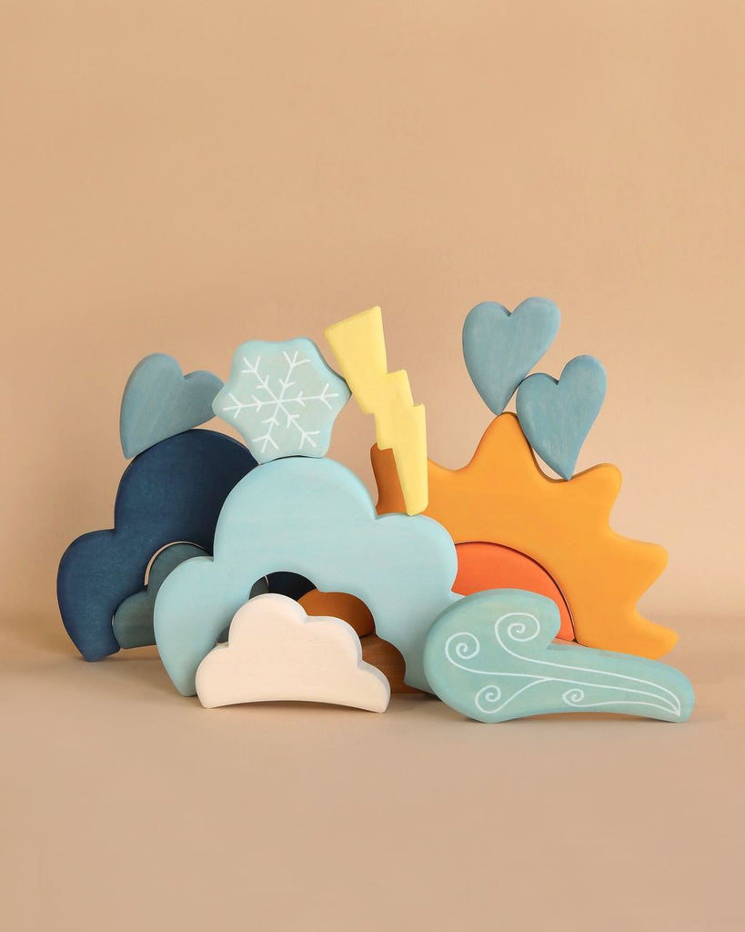 A set of colorful wooden weather-themed toys arranged on a beige background. These weather play pieces include shapes of clouds, a sun, a snowflake, hearts, a lightning bolt, and a swirling wind. Perfect for young builders, the pieces are painted in pastel shades of blue, yellow, white, and orange. Grimm's Weather Building Block Set