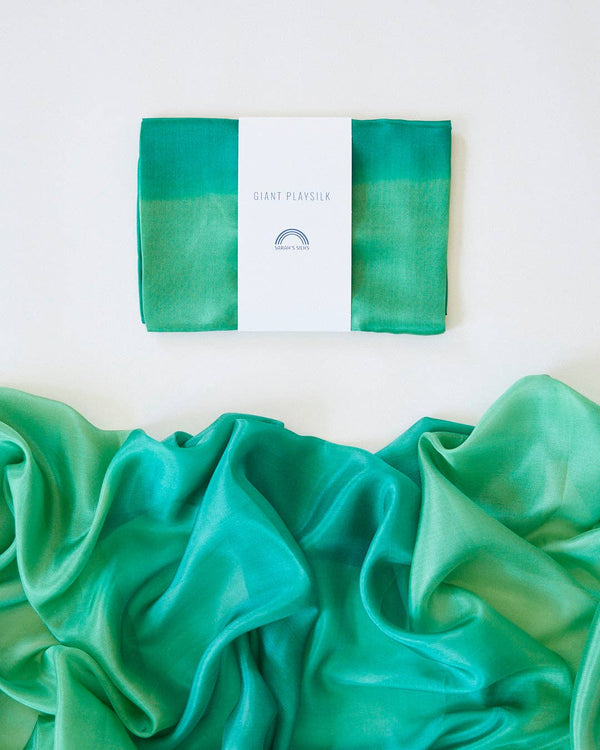 A neatly folded Sarah's Silk Giant Forest Playsilk with a minimalist label sits at the top of the image. Below it, the giant playsilk is unfolded, revealing its flowing texture and rich green hues—ideal for imaginative play that inspires creativity and open-ended adventures.