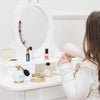 A young girl sits at a white vanity with the Vanity Star Beauty Kit, holding a pink toy hairdryer and surrounded by play makeup and accessories, her face partially reflected in the mirror.