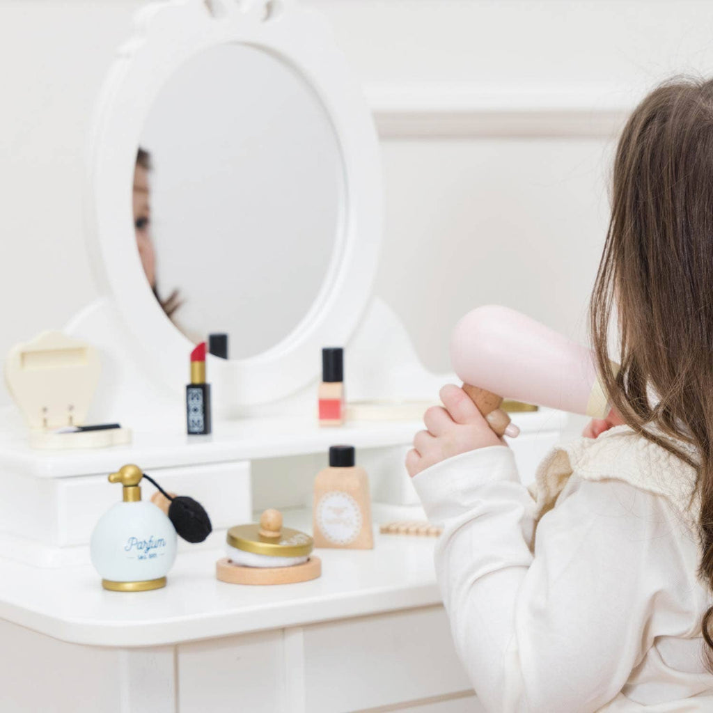 A young girl sits at a white vanity with the Vanity Star Beauty Kit, holding a pink toy hairdryer and surrounded by play makeup and accessories, her face partially reflected in the mirror.