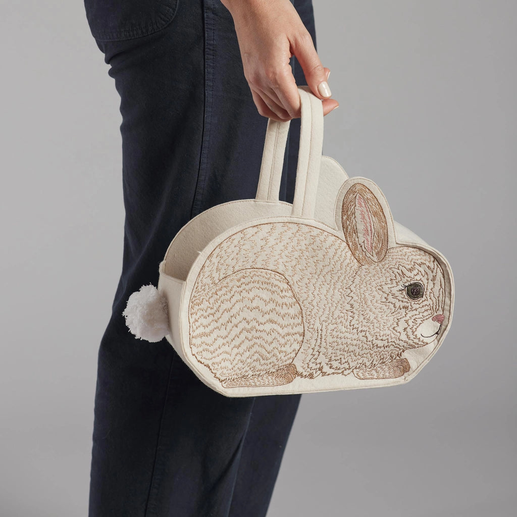 A person holding a Coral & Tusk Bunny Basket made of cream-colored fabric, featuring detailed embroidery and a fluffy tail, against a gray backdrop.