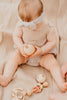 A baby in a striped romper and headband sits on a beige blanket, holding the Sabo Concept Bear Stacker - Beige. Additional wooden toys lie near the baby's legs, with the baby's face turned away from view.