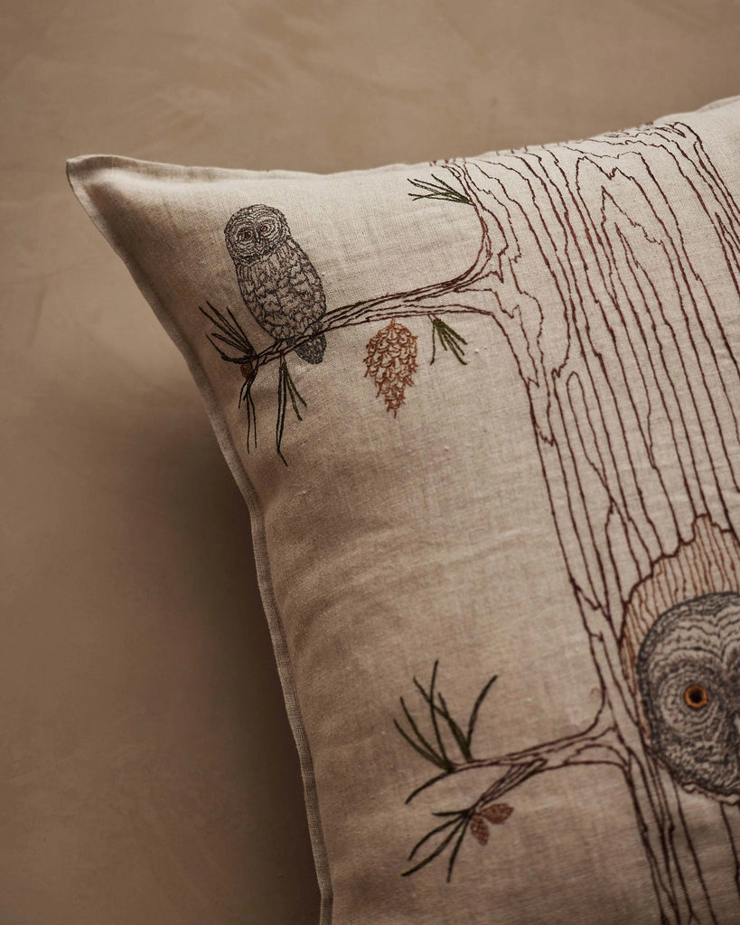 A close-up of the Coral & Tusk Owl Family Tree Pillow, showcasing embroidered owls on a tree branch, with detailed pine cone and trunk accents on a neutral background.