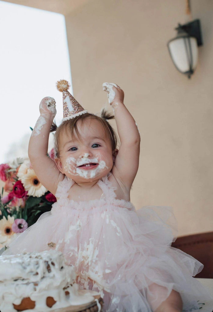 A baby wearing a pink dress and a Confetti Glitter Party Hat sits with frosting smeared on her face and hands, looking joyful with her arms raised during birthday celebrations. A partially eaten cake is in front of her, and a bouquet of flowers in the background adds charm to the photo shoot.