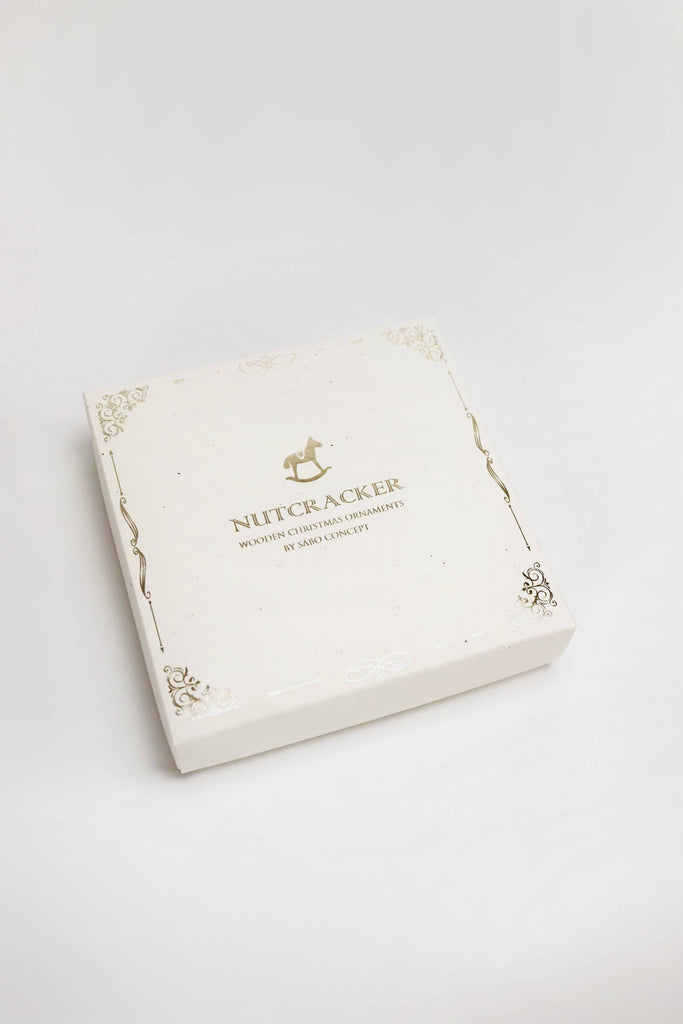 A cream-colored box with gold accents holds a hand-painted wooden Sabo Concept Christmas Ornaments set, featuring ballerina decorations, shown against a white background.