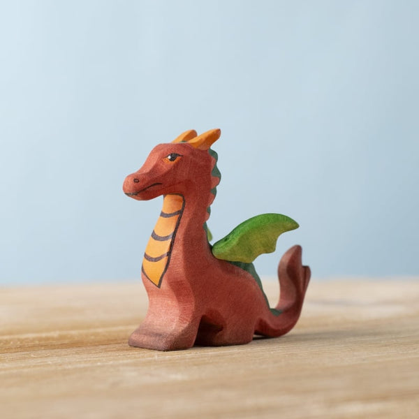 The Bumbu Handmade Wooden Baby Dragon is a small, colorful figurine with orange and green details, standing on a light wooden surface against a pale blue background—perfect for imaginative fantasy play.