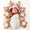 A newborn baby, wearing a hand-knit bunny hat, sleeps peacefully wrapped in a blanket with a leaf pattern, flanked by four small Cuddle + Kind Baby Fawn plush deer.